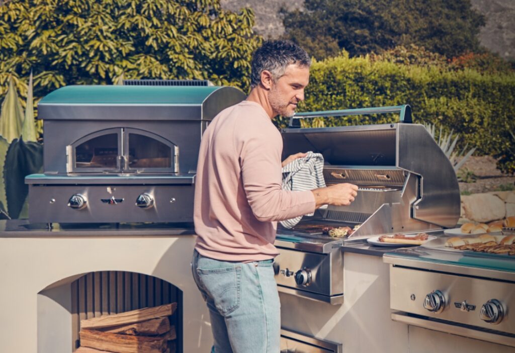 Family man cooking on a premium outdoor kitchen grill, pizza oven behind him. All available at America Home Supply in Dania Beach, Florida,